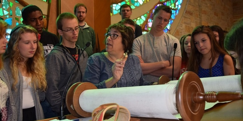 A woman reading a Torah scroll to a group of teenagers