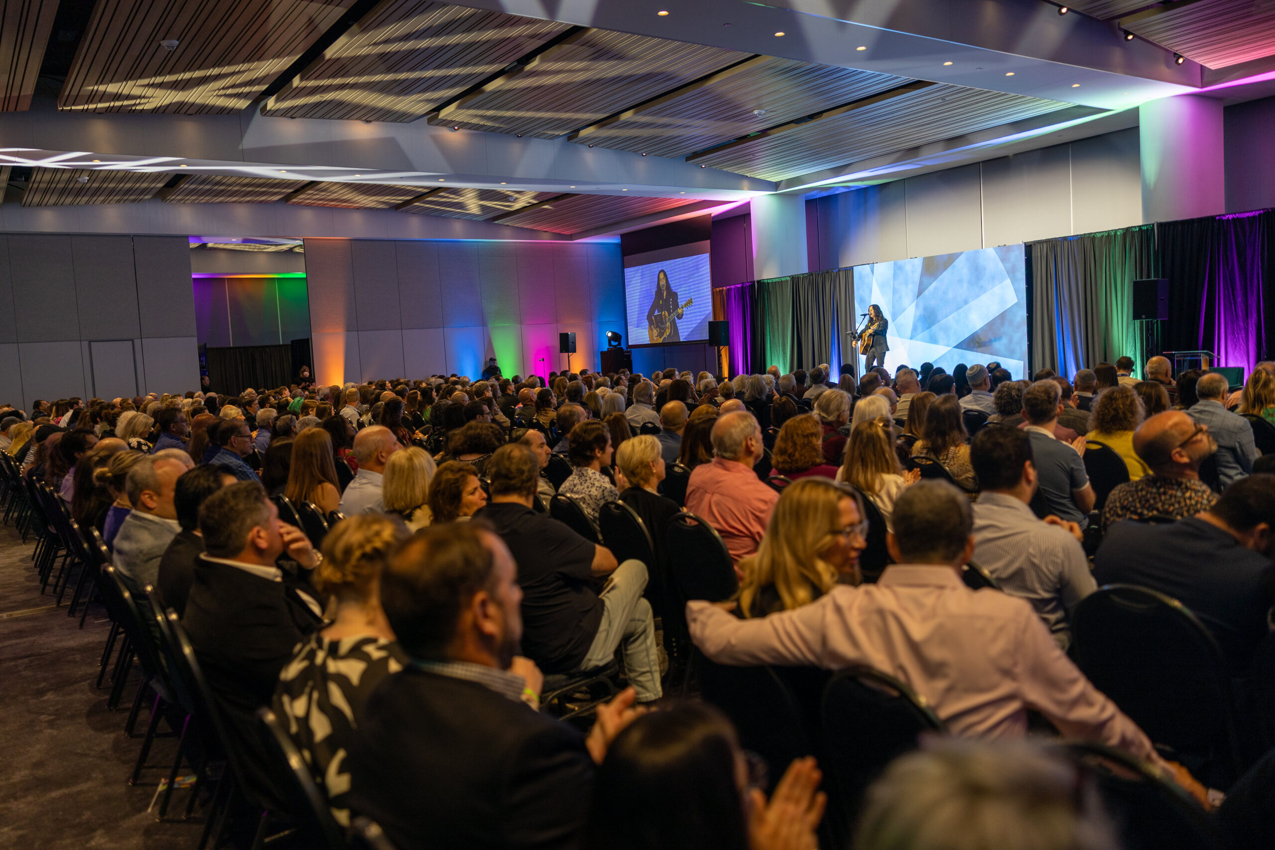 Large audience seated in a modern conference hall facing a stage where a performer stands under colorful lighting.