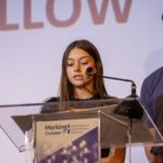 a woman speaking at a podium with man in background