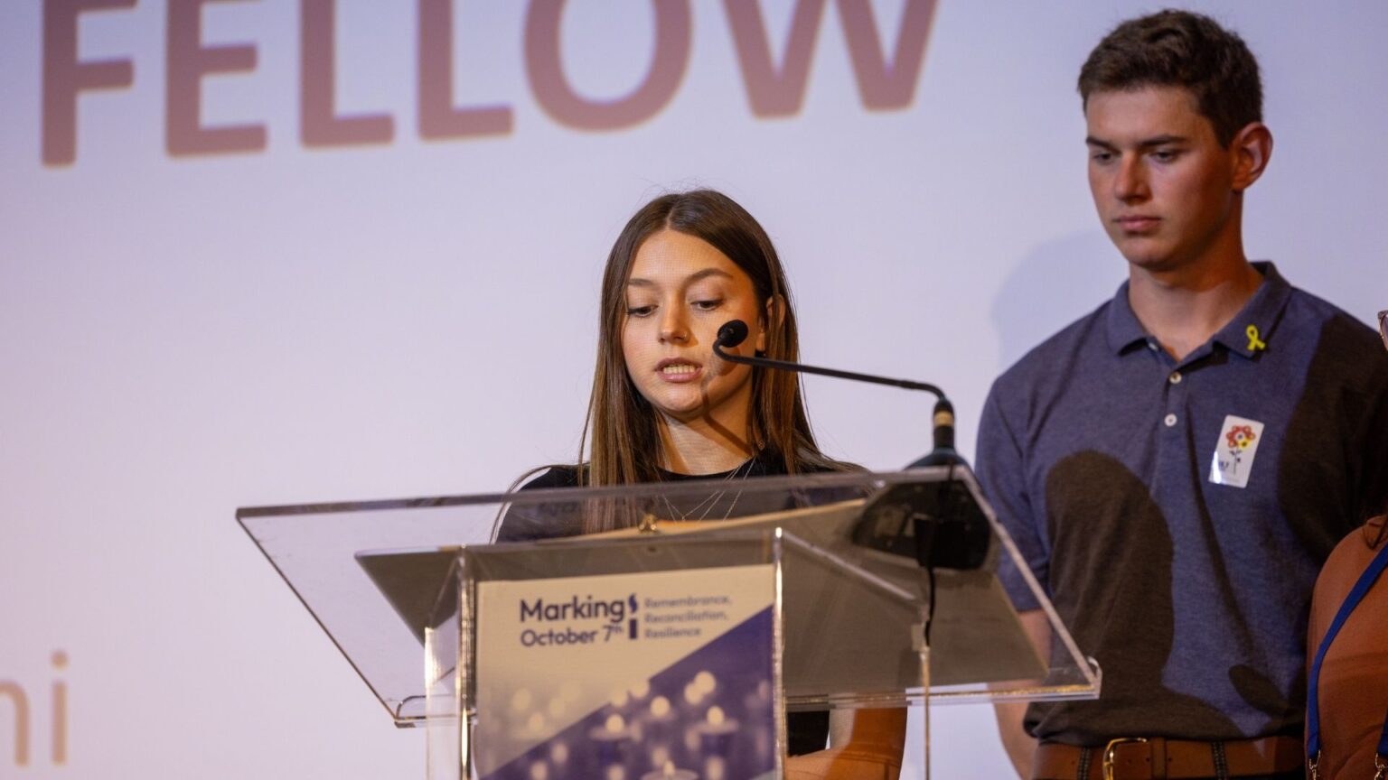 a woman speaking at a podium with man in background