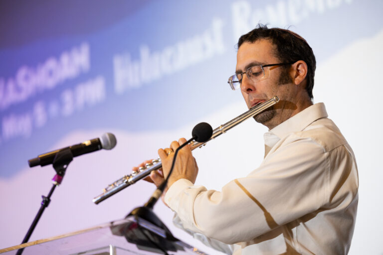 A man plays a flute at a podium with microphones, with a projected screen in the background.