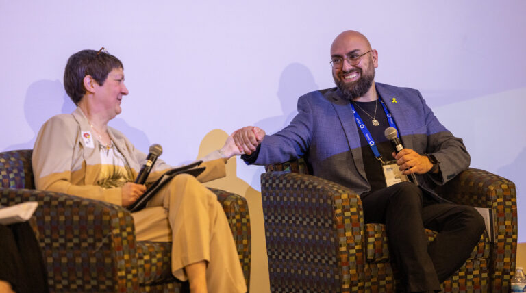 Two panelists seated in armchairs on a stage hold microphones and smile as they reach across to hold hands.